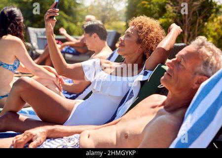 Senior Woman Using Mobile Phone Relaxing On Sun Lounger By Outdoor Pool On Summer Vacation Stock Photo