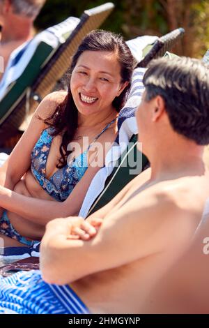 Mature Couple Talking On Loungers Relaxing By Swimming Pool On Summer Vacation Stock Photo