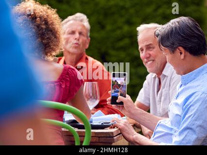 Man Taking Photo As Group Of Friends Talking Around Table At Summer Dinner Party In Garden At Home Stock Photo