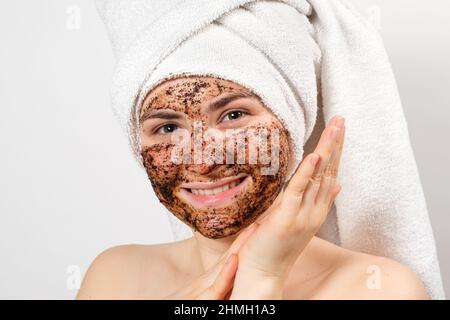 A young woman with a coffee mask on her face and a bath towel on her head smiles and looks into the camera on a white background. Stock Photo