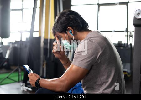 focus on hand, young indian body builder with medical face mask cleaning sweat while using mobile phone at gym - concept of covid-19 coronavirus Stock Photo
