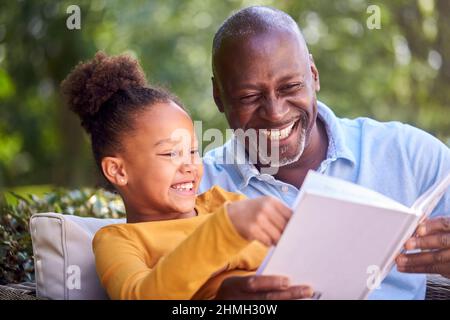 Grandfather Sitting Outdoors With Granddaughter At Home Reading Book Together Stock Photo