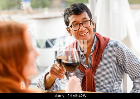 Positive ethnic male clinking glass of red wine with female friend and looking away at table in street restaurant Stock Photo