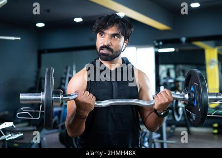 Young Indian bodybuilder busy doing biceps workout by lifting weight using barbells at gym - concept of musclebuilder exercising, healthy lifestyle Stock Photo