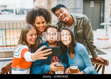 Group of positive multiracial friends making funny faces while taking self portrait on smartphone at cafe table Stock Photo