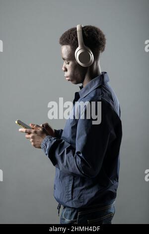 Side view of confident young African American male millennial in trendy outfit and headphones using mobile phone while listening to music against gray Stock Photo