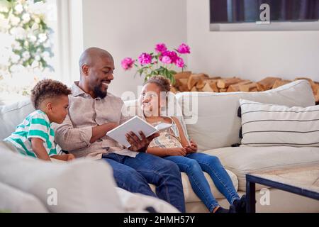 Father Sitting On Sofa With Children At Home Reading Book Together Stock Photo