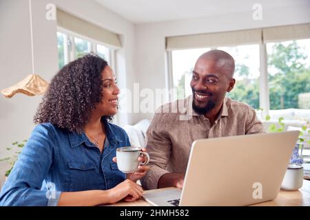 Couple Sitting Around Table At Home Shopping Or Booking Holiday On Laptop Stock Photo