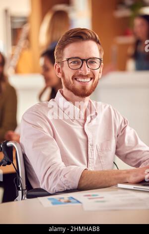 Portrait Of Businessman In Wheelchair Sitting At Desk Working On Laptop In Busy Office Stock Photo