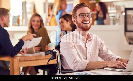 Laughing Businessman In Wheelchair Sitting At Desk Working On Laptop In Busy Office Stock Photo