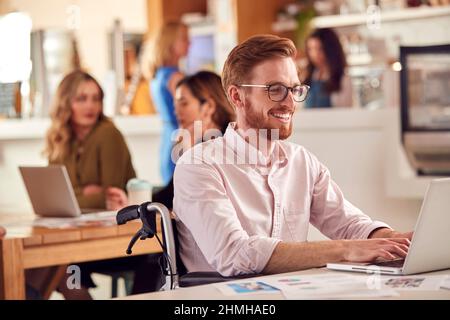 Businessman In Wheelchair Sitting At Desk Working On Laptop In Busy Office Stock Photo