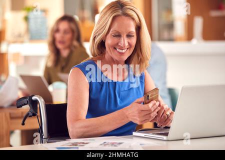 Mature Businesswoman In Wheelchair Sitting At Desk Working On Laptop In Busy Office Stock Photo