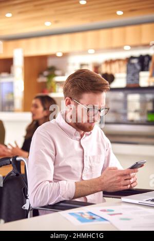 Businessman In Wheelchair With Mobile Phone Sitting At Desk Working On Laptop In Busy Office Stock Photo