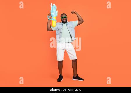 Photo of strong guy dressed jeans waistcoat dark glasses shooting water gun rising fist isolated orange color background Stock Photo