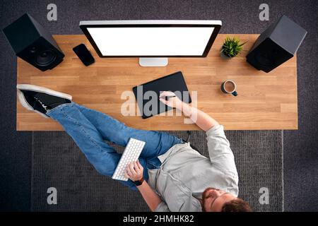 Overhead View Of Casual Male Graphic Designer At Computer With Feet On Desk And Keyboard On Lap Stock Photo