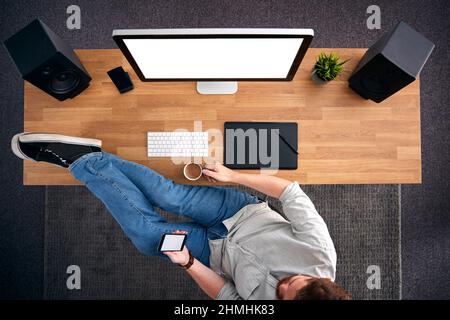 Overhead View Of Male Graphic Designer At Computer With Feet On Desk Looking At Mobile Phone Stock Photo