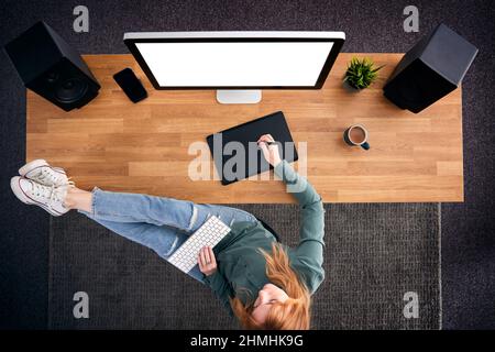 Overhead View Of Casual Female Graphic Designer At Computer With Feet On Desk And Keyboard On Lap Stock Photo