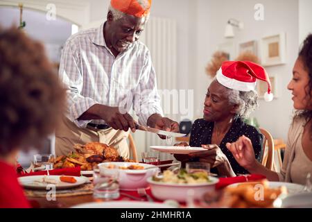 Grandfather Carving And Serving As Multi Generation Family Enjoy Eating Christmas Meal At Home Stock Photo
