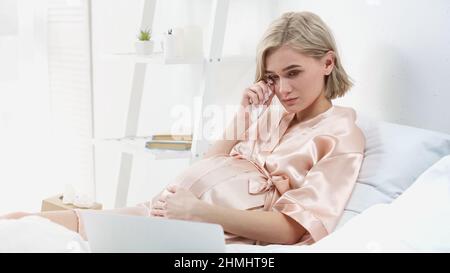 sentimental blonde pregnant woman crying and wiping tear while watching movie on laptop in bedroom Stock Photo