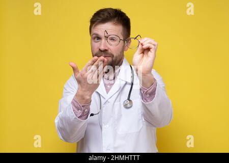 Doctor in a white coat smiles in surprise, makes a gesture with hand and looks with one eye through the lens of glasses.  Stock Photo