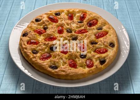 Apulian focaccia, typical italian Bari pizza with cherry tomatoes and black olives on top. In white plate on light blue wooden table Stock Photo