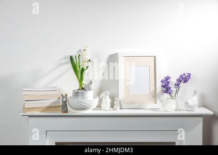 White hyacinth in a large porcelain bowl, books, figurines of hares and a bird, an empty photo frame are on the fireplace against the white wall. Layo Stock Photo