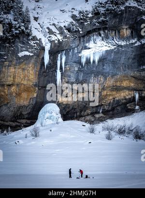 people ice fishing in front of an impressive rock wall with icicles hanging from it and forming an ice tower below. Stock Photo