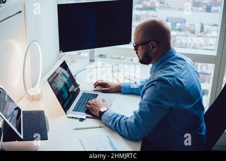 Serious male employee typing on netbook sitting at table with computer and tablet Stock Photo