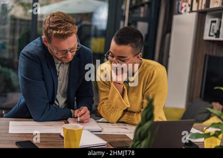 Two diverse natural colleagues in casual office or home office doing paperwork working together teaching, coaching or tutoring each other one another Stock Photo