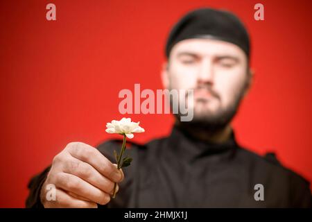 A male security guard in a black uniform and a cap holds a flower in his hand. Bodyguard. Defender of the law and order. Military. Mercenary. Handsome Stock Photo