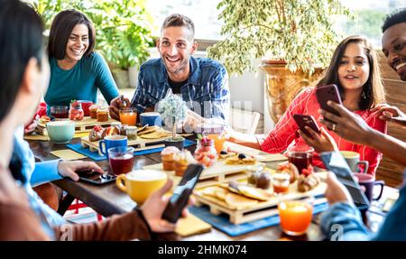 Multiracial friends interacting with mobile phone at coffee bar - People having fun at cafeteria on morning brunch time - Life style concept with happ Stock Photo