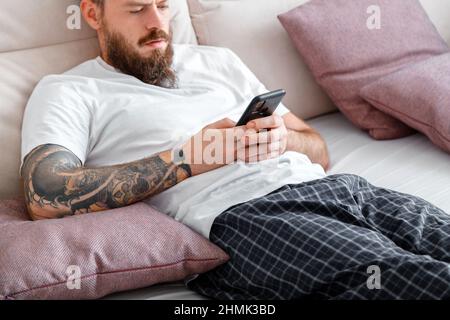 Man in pajamas using smartphone while lying in bed. Young caucasian bearded brutal with tattoos man writes message on mobile phone or surfing in Stock Photo