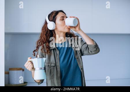 Curly-haired young woman with loose hairstyle dances to loud music in wireless headphones with coffee in cup and coffeepot Stock Photo
