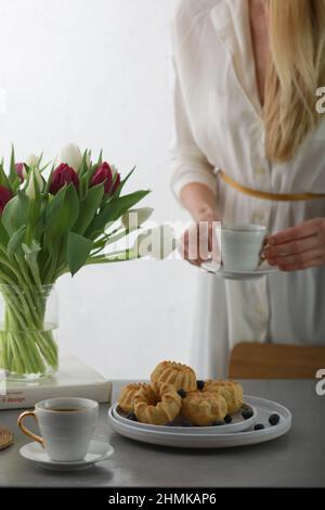 Mini bundt cakes with fruits on plate, two cups of coffee and bunch tulip flowers in a vase. Girl holding cup of coffee. Stock Photo