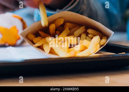Young African-America man takes delicious fresh french fries from paper container at table in modern cafe extreme close view Stock Photo