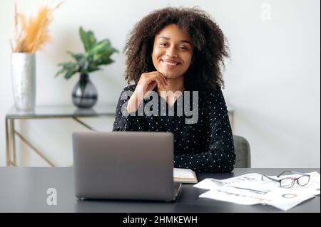 Portrait of attractive african american successful business woman, manager, business consultant, trader, dressed in formal stylish shirt, sitting at workplace with laptop, looking at camera, smiling Stock Photo