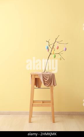 Vase with tree branch and Easter eggs on wooden stool against color wall Stock Photo