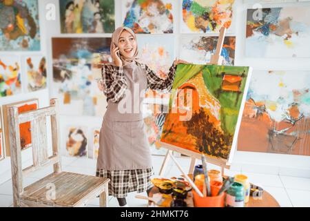 muslim asian female artist painting on canvas and talking to phone Stock Photo