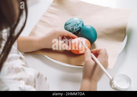 Cropped portrait of girl hand, painting multi coloring Easter hens eggs on brown paper plate table with paintbrush. Making art design white patterns o Stock Photo