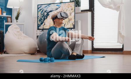 Retired woman meditating in lotus position with vr glasses on yoga mat. Pensioner using virtual reality headset and doing meditation to relax. Senior person wearing goggles to meditate. Stock Photo