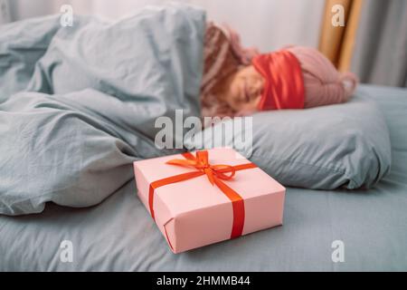 Woman in red sleep mask sleeping in bed near paper gift box with red satin ribbon decor. Christmas, New Year, Valentine's Day and birthday concept Stock Photo