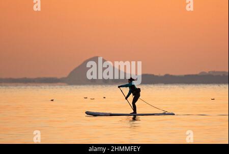 Portobello, Edinburgh, Scotland, UK. 11th February 2022. Freezing temperatures at sunrise with minus 2 degrees for  those brave enough to venture out onto the Firth of Forth. Pictured: A female paddleboarderwith Berwick Law in East Lothian in the background takes exercise at dawn. Stock Photo