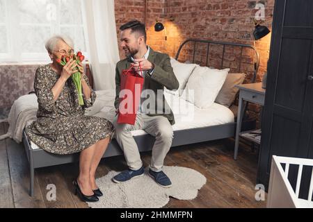 wide shot of a caucasian middle-aged man giving his mother beautiful red tulips and a present for Mother's Day . High quality photo Stock Photo