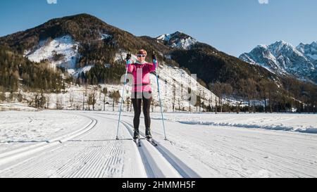 Smiling middle aged female recreational cross country skier enjoying nature activity, gliding in trails cut into the snow, tracking shot. Stock Photo