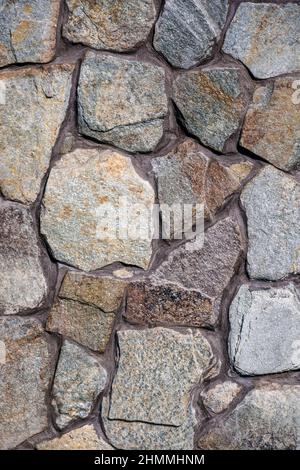 Concrete wall trimmed with wild multi-colored stone. The seams are painted with brown paint. Country style. Close-up. Selective focus. Stock Photo