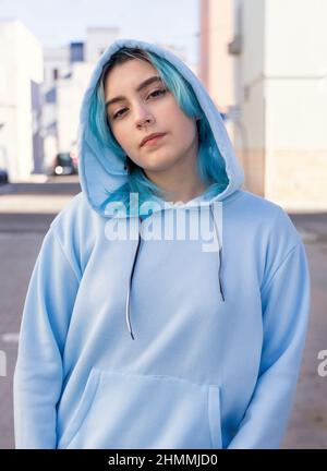 Teenager wearing light blue oversize hoodie and jeans looking into the camera. Blue haired teen girl stays outdoor on city street. Clothing mockup Stock Photo