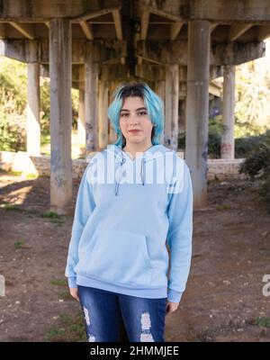 Smiling Teenager wearing light blue oversize hoodie and jeans stays under bridge . Blue haired teen girl stays outdoors against bridge pillars. Clothi Stock Photo