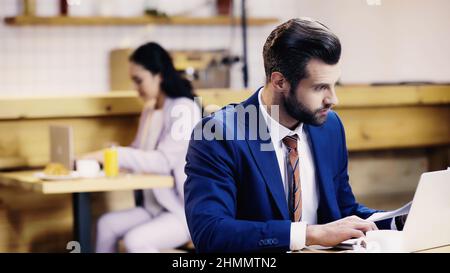 surprised businessman looking at documents near laptop and blurred woman in cafe Stock Photo