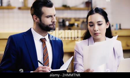 bearded businessman writing in notebook near asian businesswoman with documents in cafe Stock Photo