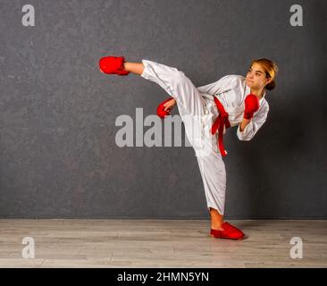 A young karateka girl in a white kimono and red competition outfit trains and performs a set of exercises against a gray wall Stock Photo